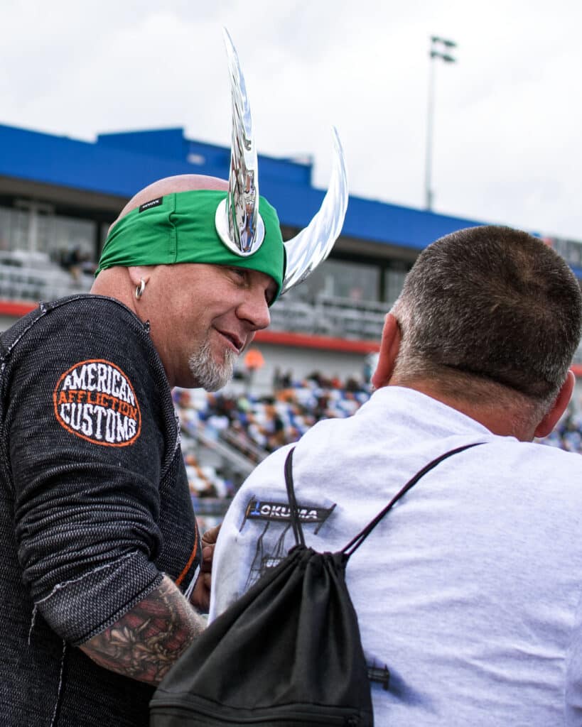 two nhra drag racing fans talk to each other, one with a white shirt and another with a black shirt, green headband and makeshift horns