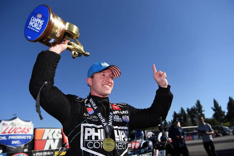 Justin Ashley, NHRA Top Fuel driver celebrating a win with a trophy in hand and black racing suit on