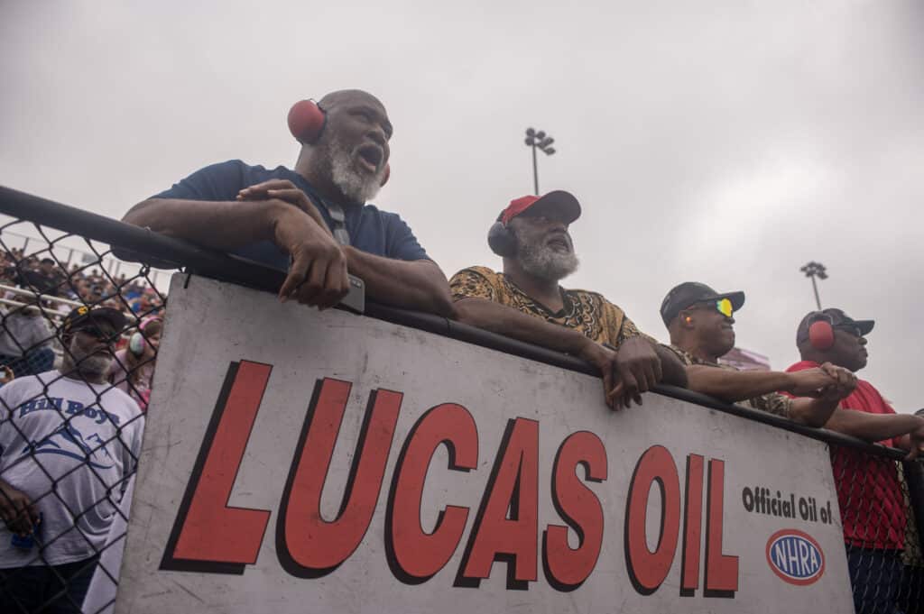 four fans lean against a fence to watch an nhra drag race with a lucas oil sign in front of them
